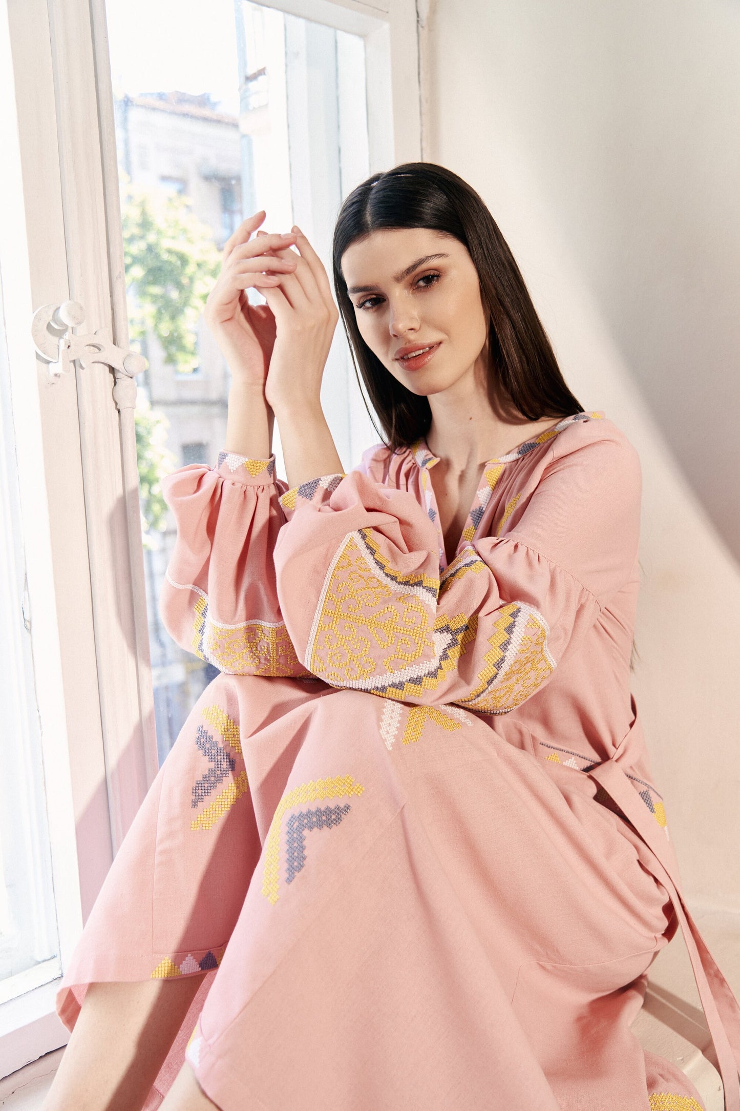 Woman wearing pink linen embroidered Vyshyvanka dress with boho design, belt, and pockets, sitting by a window
