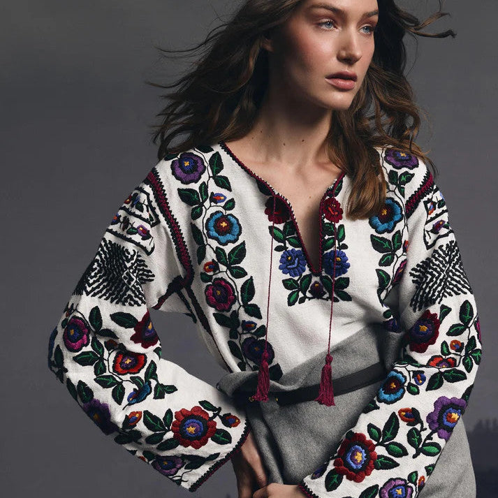 Woman wearing a white blouse with colorful floral embroidery, paired with a grey skirt, posing against a dark, moody background.