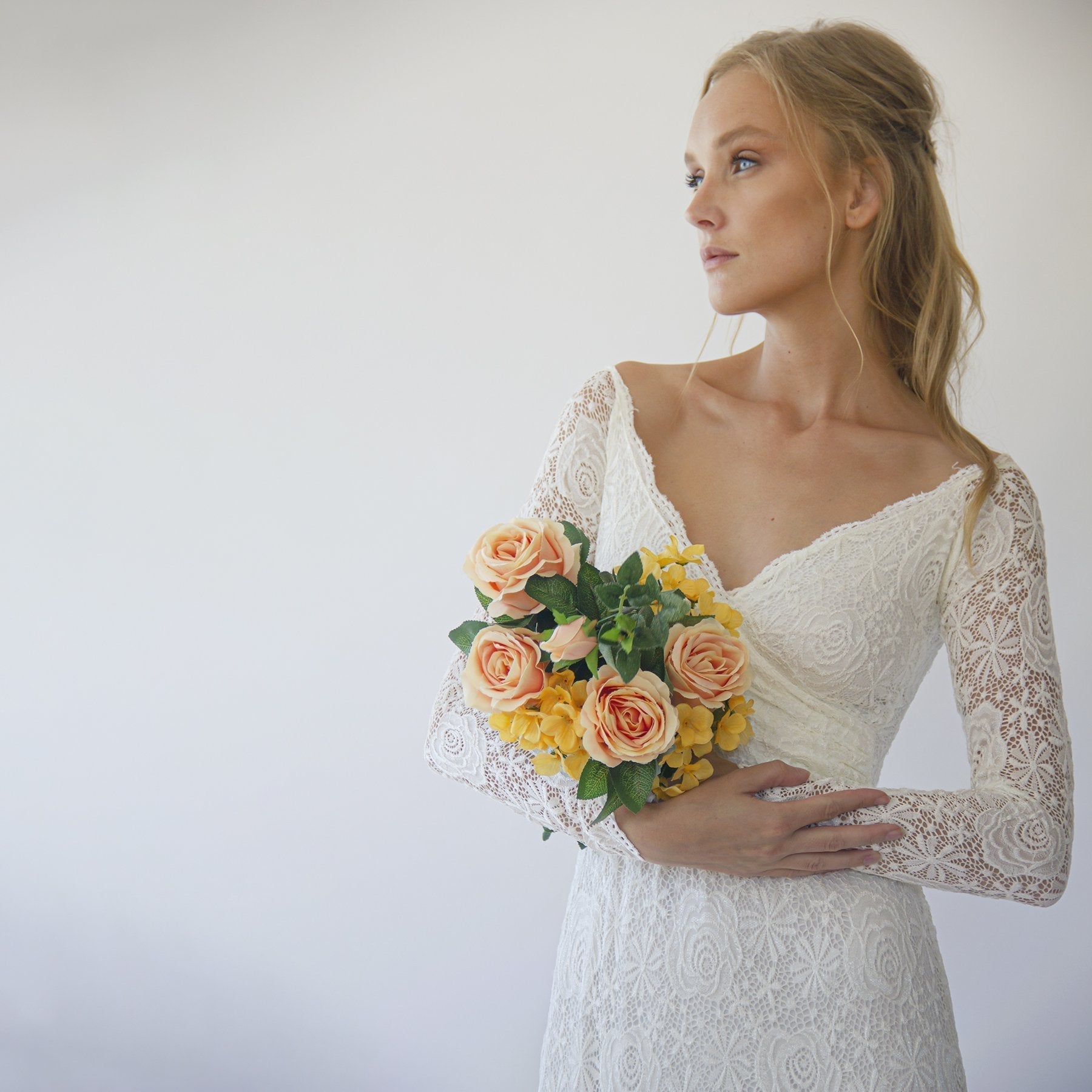 Bride in lace wedding dress holding bouquet of roses and yellow flowers on white background.