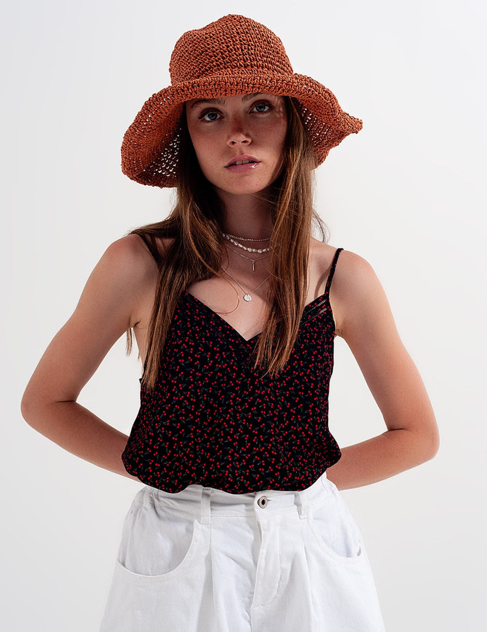 Woman wearing a black floral top, white pants, and a brown woven hat against a white background.