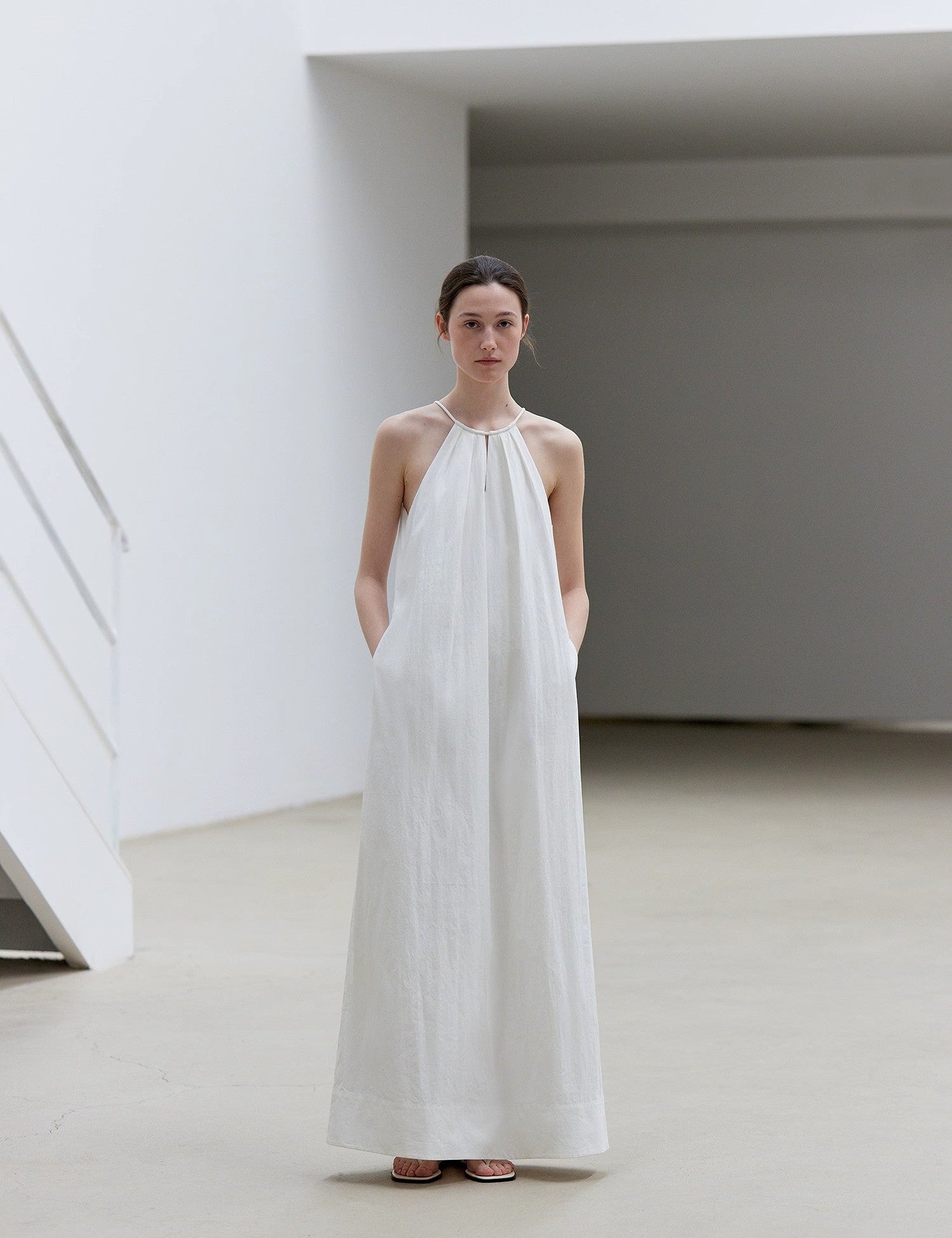 Model wearing white Veridia Halter-Pleat Maxi Dress, showing halter tie, V-shaped front cut-out, and pleated skirt with elegant flow.