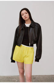Person wearing a black leather jacket with yellow shorts, standing against a plain wall.