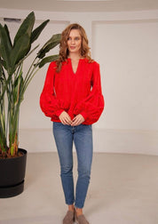 Woman wearing a red linen embroidered blouse in Ukrainian vyshyvanka boho style, paired with jeans, beside a potted plant.