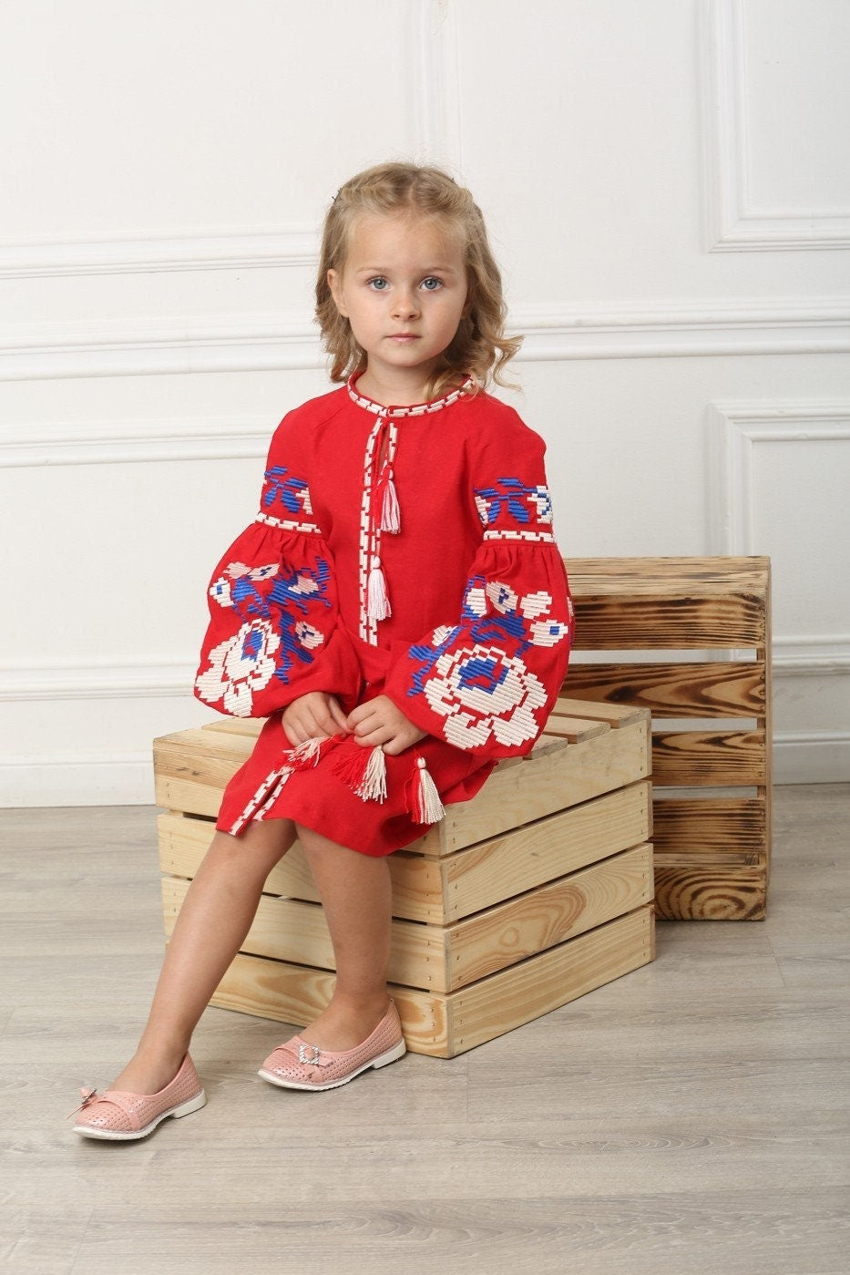 Girl wearing 2025 red linen embroidered Ukrainian vyshyvanka dress with floral cross-stitch, sitting on wooden boxes.