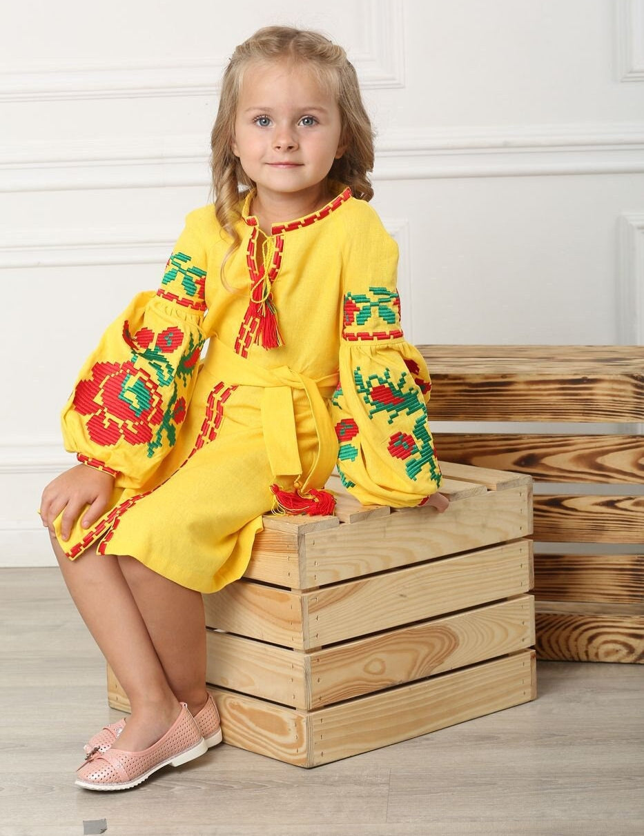 Young girl in yellow Ukrainian embroidered dress with floral cross-stitch, sitting on wooden box, showcasing traditional festive attire.