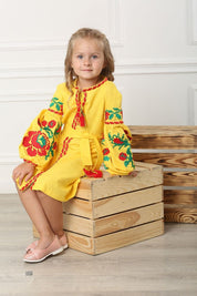 Young girl in yellow Ukrainian embroidered dress with floral cross-stitch, sitting on wooden box, showcasing traditional festive attire.
