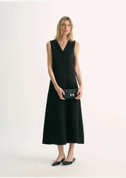 Model wearing black Aurevae Cupro-Linen A-Line Dress with V-neck and sleeveless design, paired with a clutch and flats.