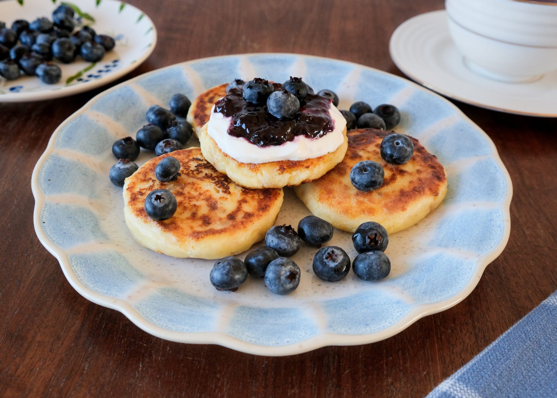 Plate of Ukrainian syrnyky cheese pancakes topped with cream and blueberries on a wooden table.