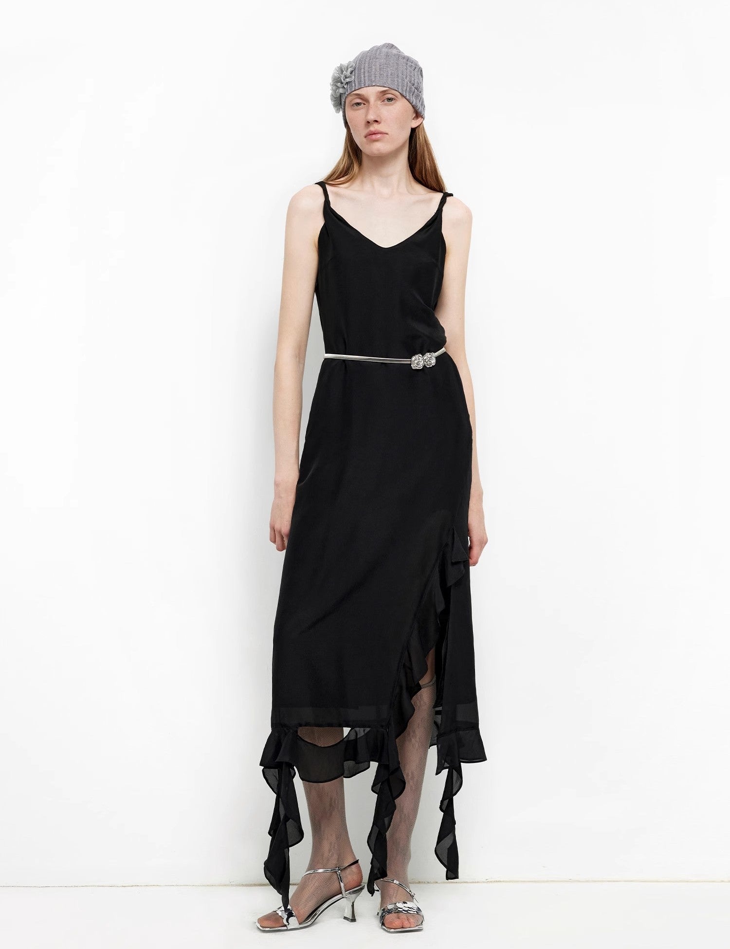 Woman wearing a black draped dress with twist straps and a subtle A-line skirt, accessorized with a grey beanie, belt, and silver heels.