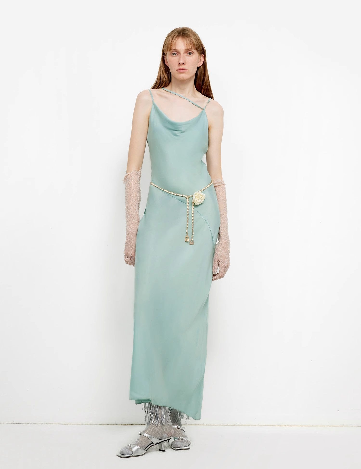 Elegant woman wearing a light blue satin gown with a cowl neckline and metallic belt, standing against a white background.