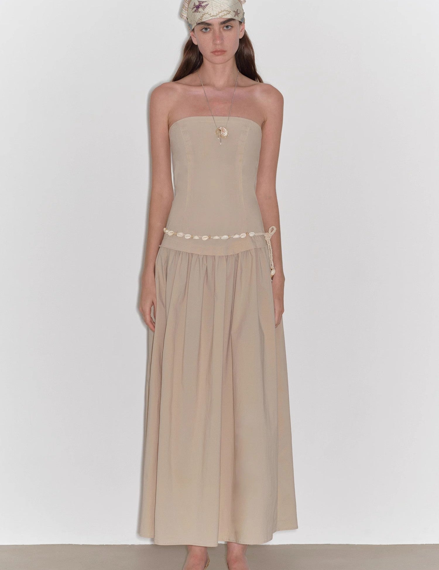 Model wearing a strapless beige A-line dress with a fitted bodice and a flowy skirt, standing in a neutral background.