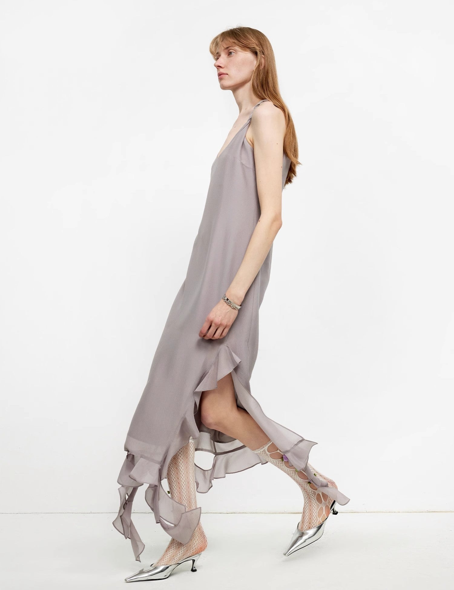 Side view of woman wearing a flowing, taupe maxi dress with ruffled hem, paired with metallic pointed-toe shoes.