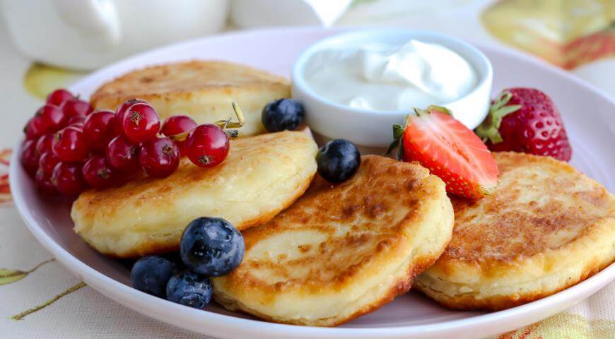 Plate of homemade Ukrainian syrnyky cheese pancakes with berries and sour cream dip, served on a white plate.