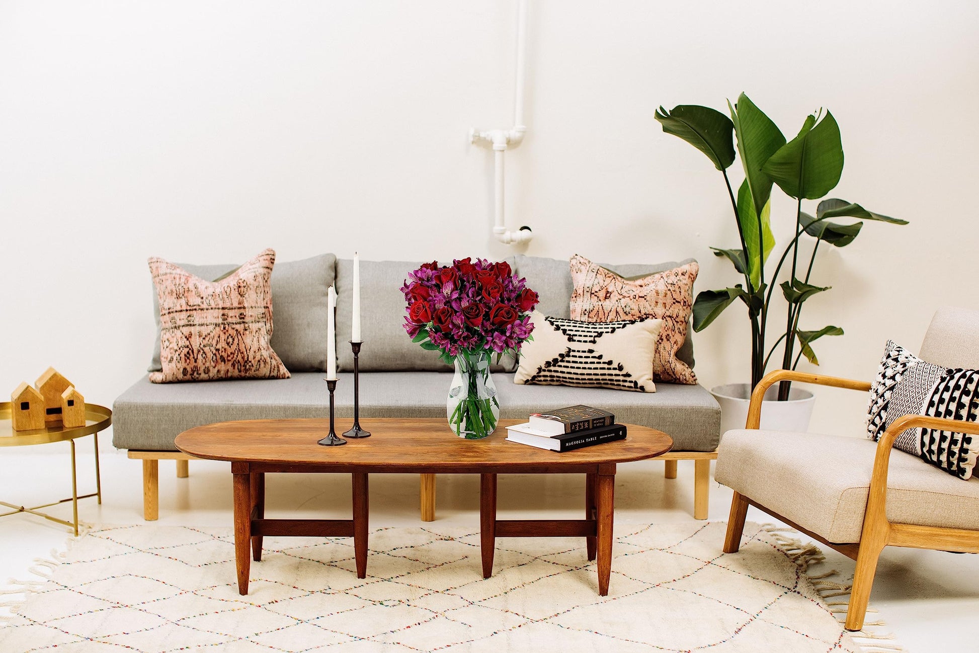 Modern living room with gray sofa, wooden table, and vibrant flower bouquet in glass vase, featuring cozy pillows and a green plant.