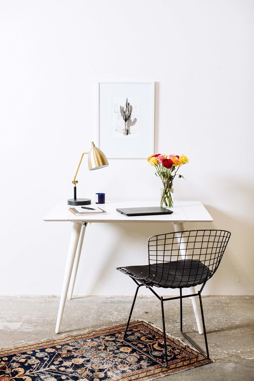 Stylish workspace with a gold lamp, white desk, laptop, and rainbow roses in a vase, complemented by a black chair and patterned rug.