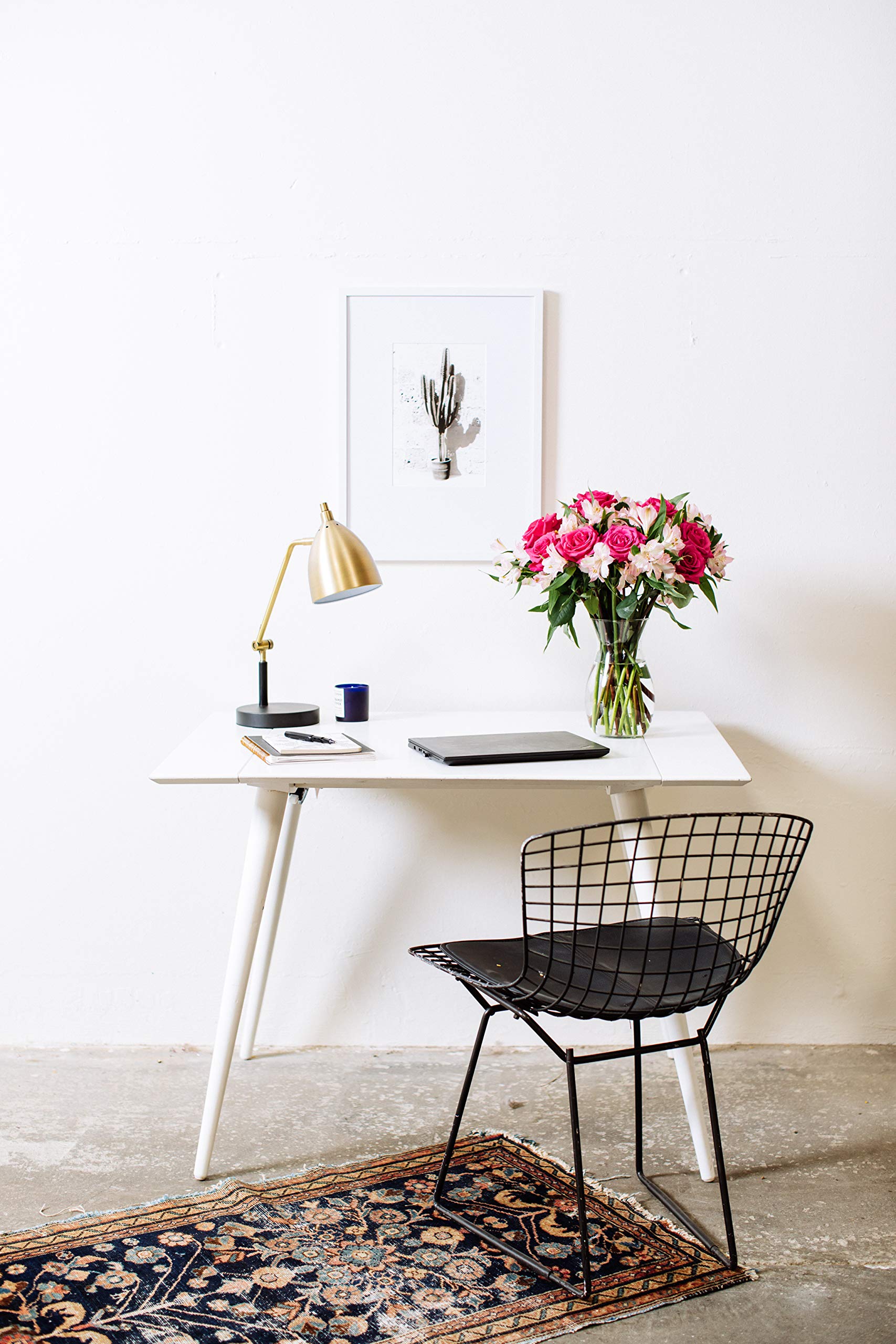 Modern workspace with a bouquet of pink roses and alstroemeria in a glass vase on a white desk, featuring a gold lamp and a framed cactus print.