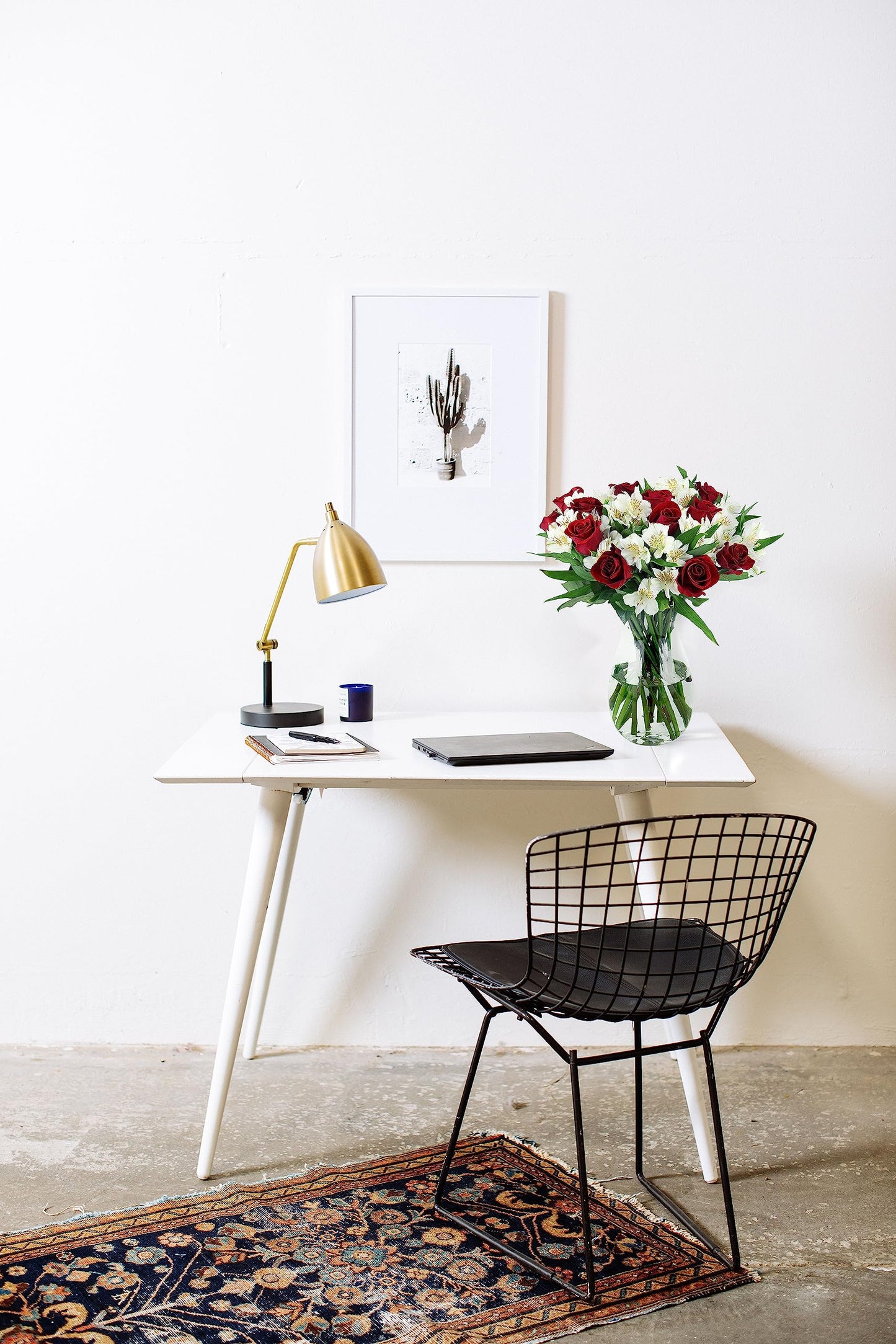 Modern workspace with a sleek white desk, gold lamp, laptop, and a vase of fresh red and white flowers against a minimalist decor background.