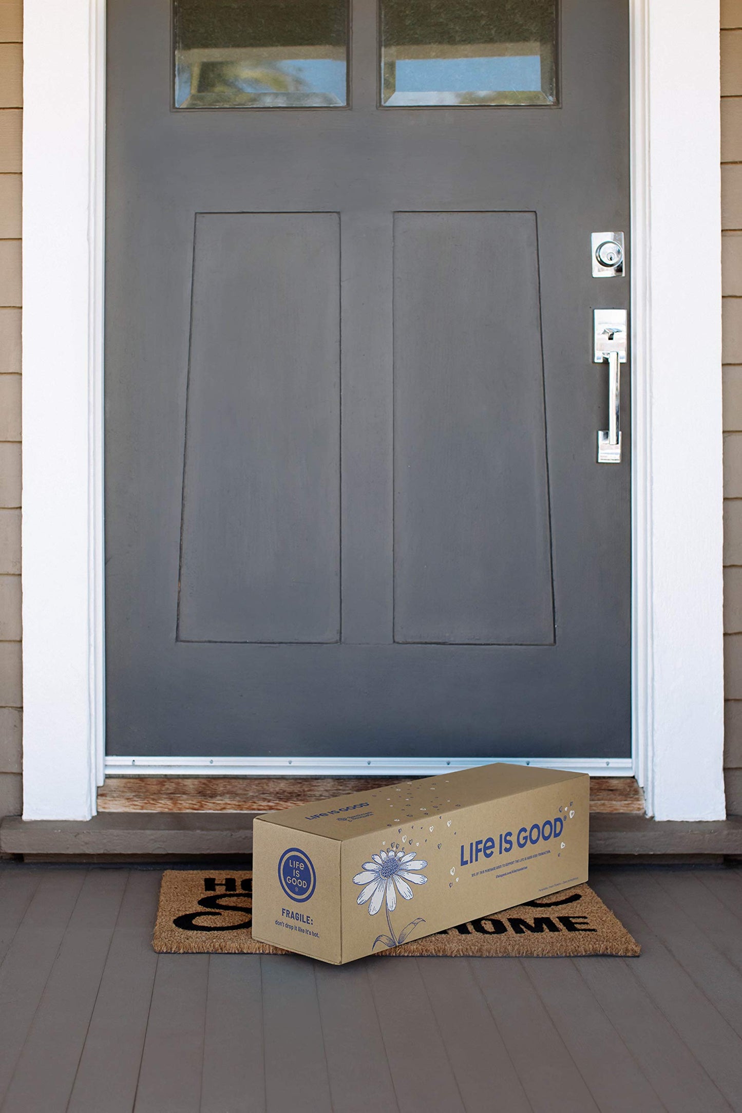 Delivery box labeled "Life is Good" on a welcome mat in front of a gray door.