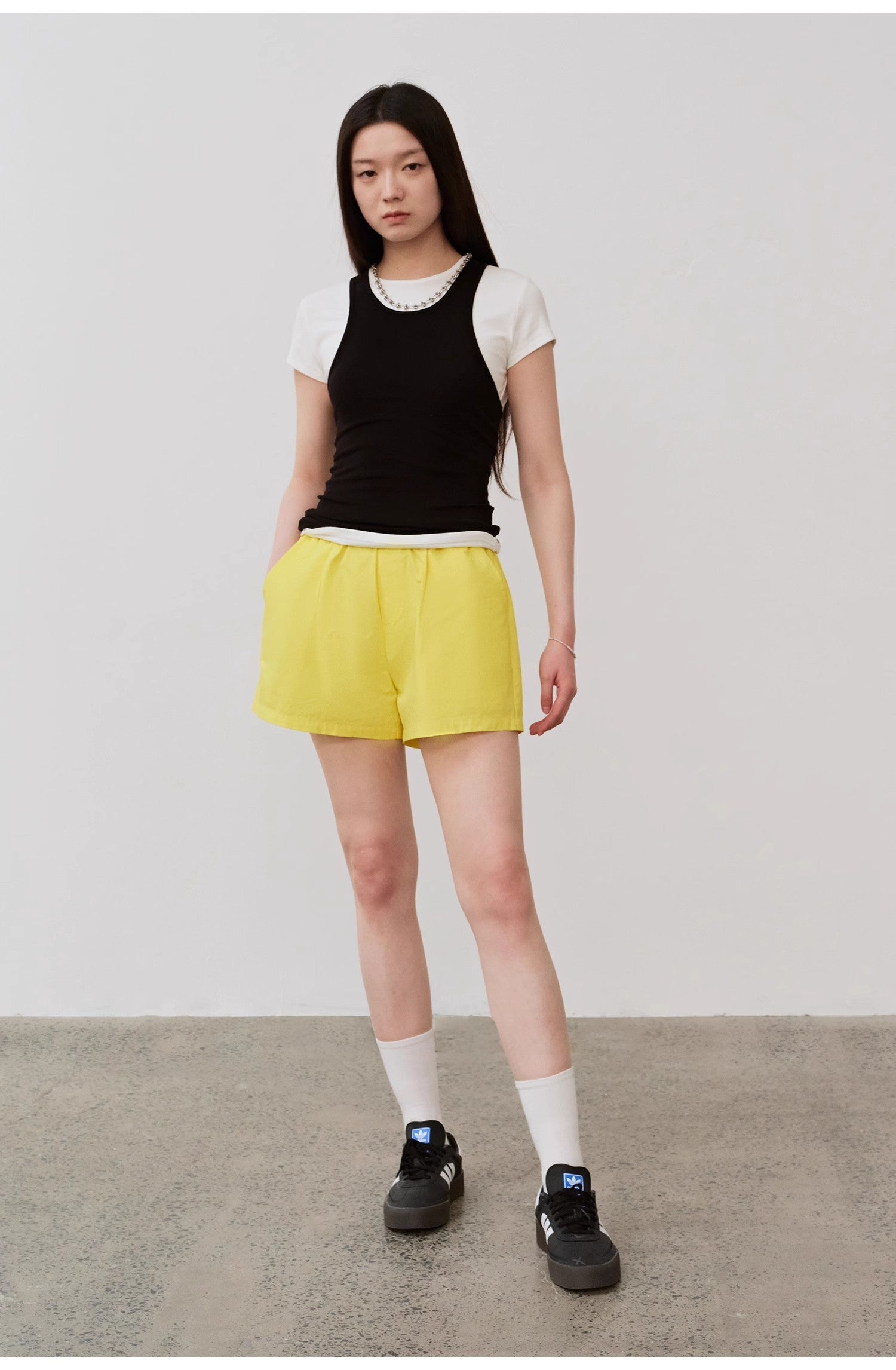 Model wearing black camisole over white top with yellow shorts and black sneakers.
