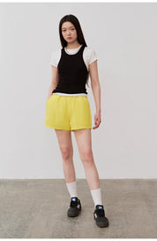 Model wearing black camisole over white top with yellow shorts and black sneakers.