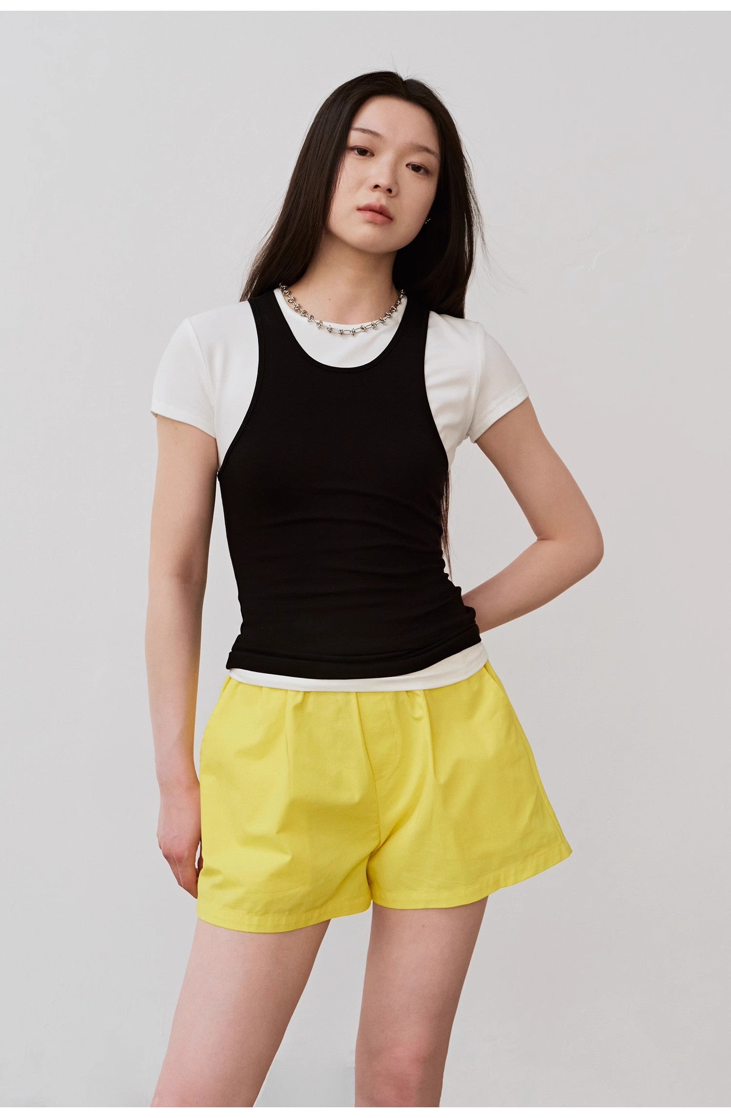 Woman wearing layered black camisole over white shirt with yellow shorts, showcasing a casual summer outfit.
