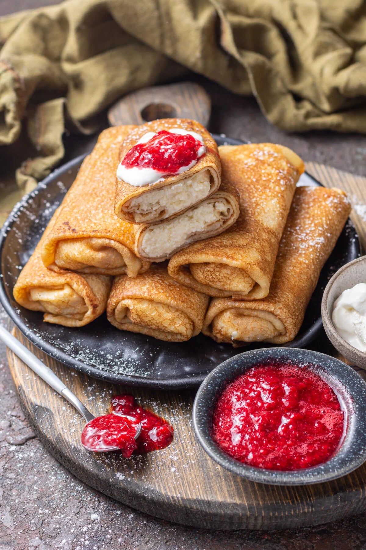 Homemade Ukrainian crepes with fillings, served on a black plate with red berry sauce and cream, showcasing a traditional savory dish.