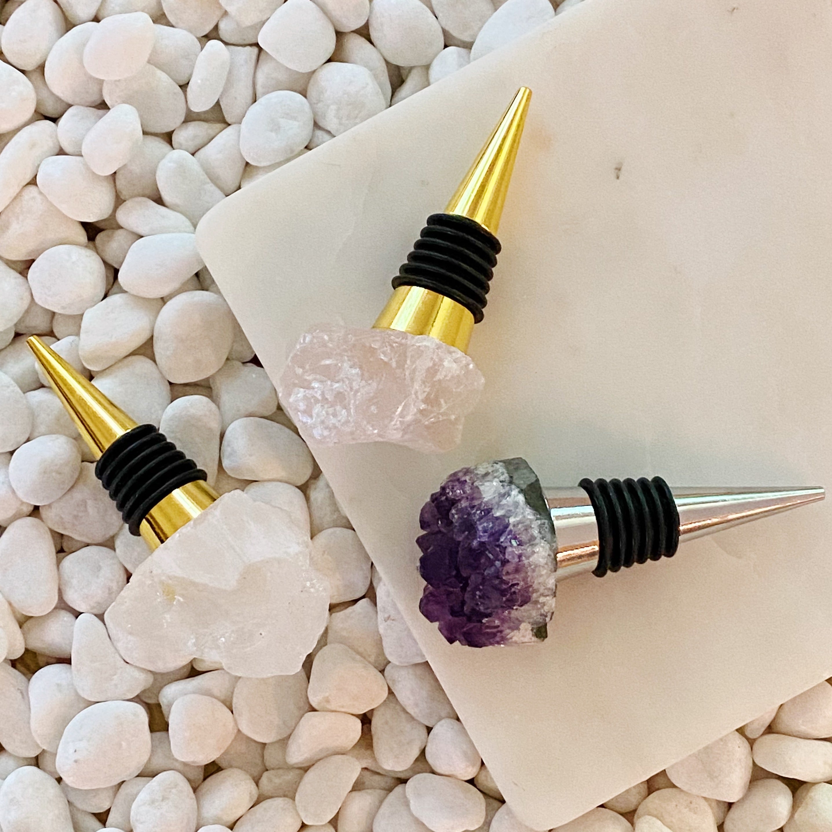 Elegantly crafted wine stoppers with natural-cut semi-precious stones on a bed of white stones, perfect for home or gifts.