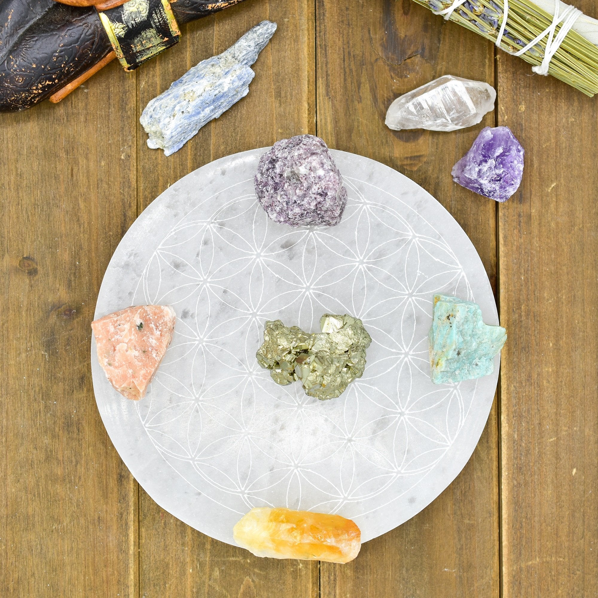 Round selenite charging plate with engraved Flower of Life symbol surrounded by assorted crystals on wooden surface