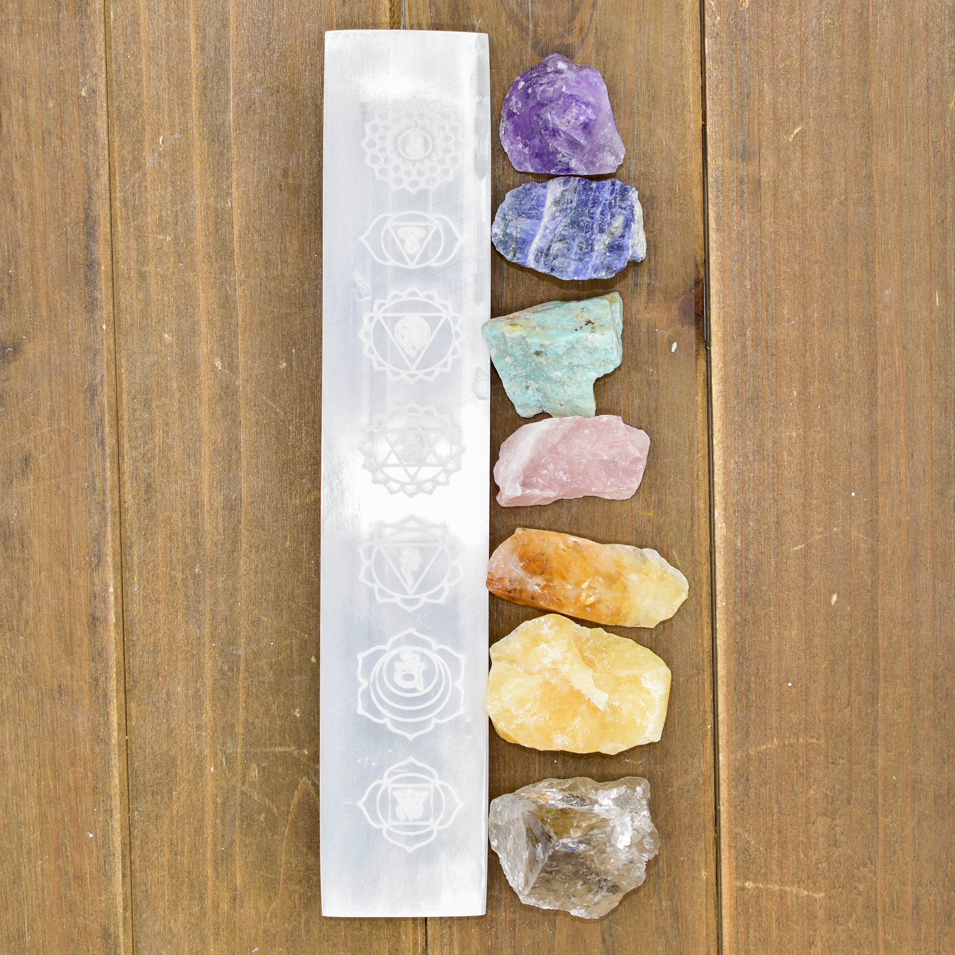 7 Chakra Selenite Charging Bar with engraved chakra symbols, surrounded by colorful healing crystals, on wooden surface.