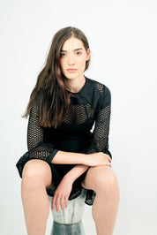 Woman wearing black Lixell Mesh-Panel Dress with airy texture and geometric design, sitting on a stool in a minimalist setting.