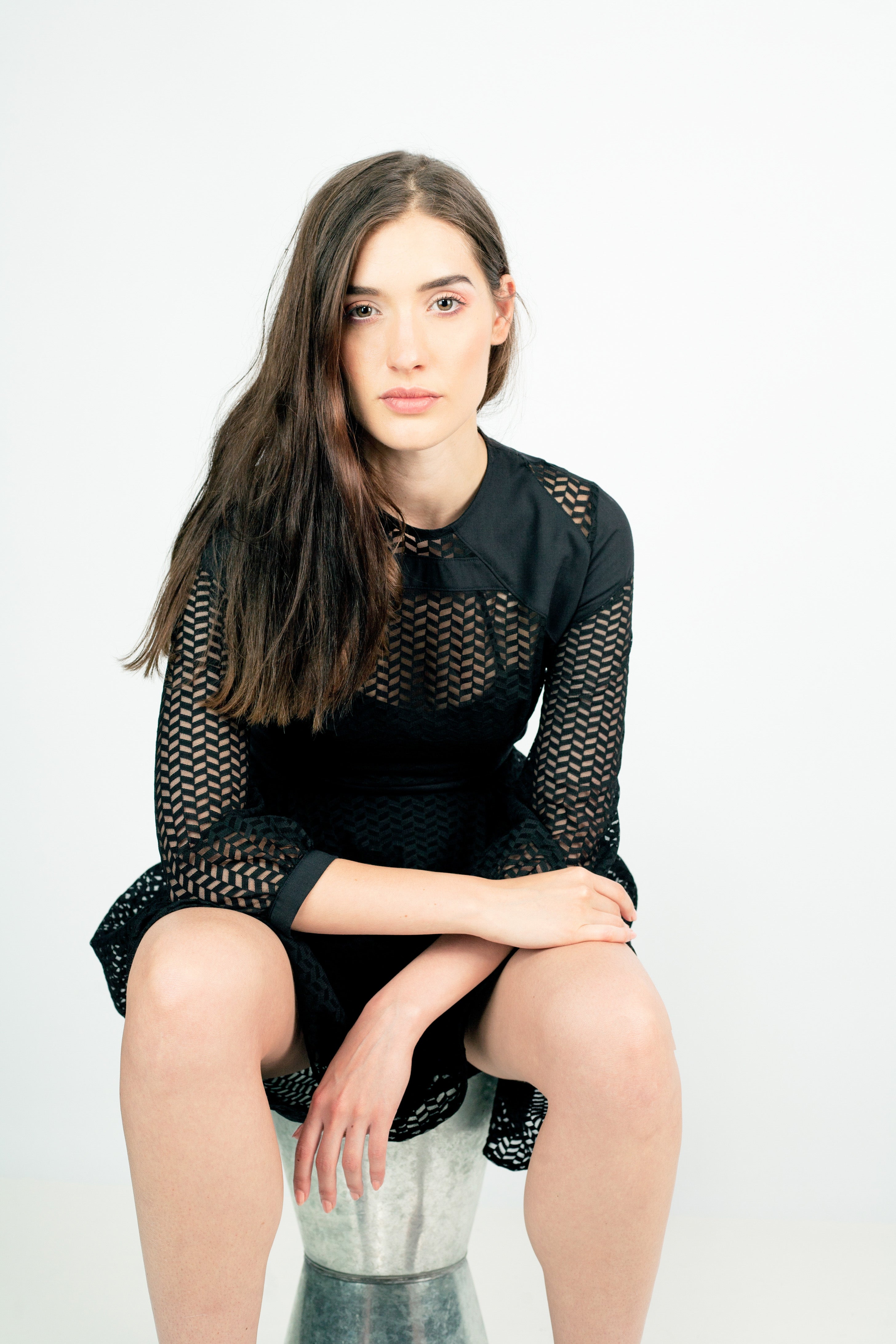 Woman wearing black Lixell Mesh-Panel Dress with airy texture and geometric design, sitting on a stool in a minimalist setting.