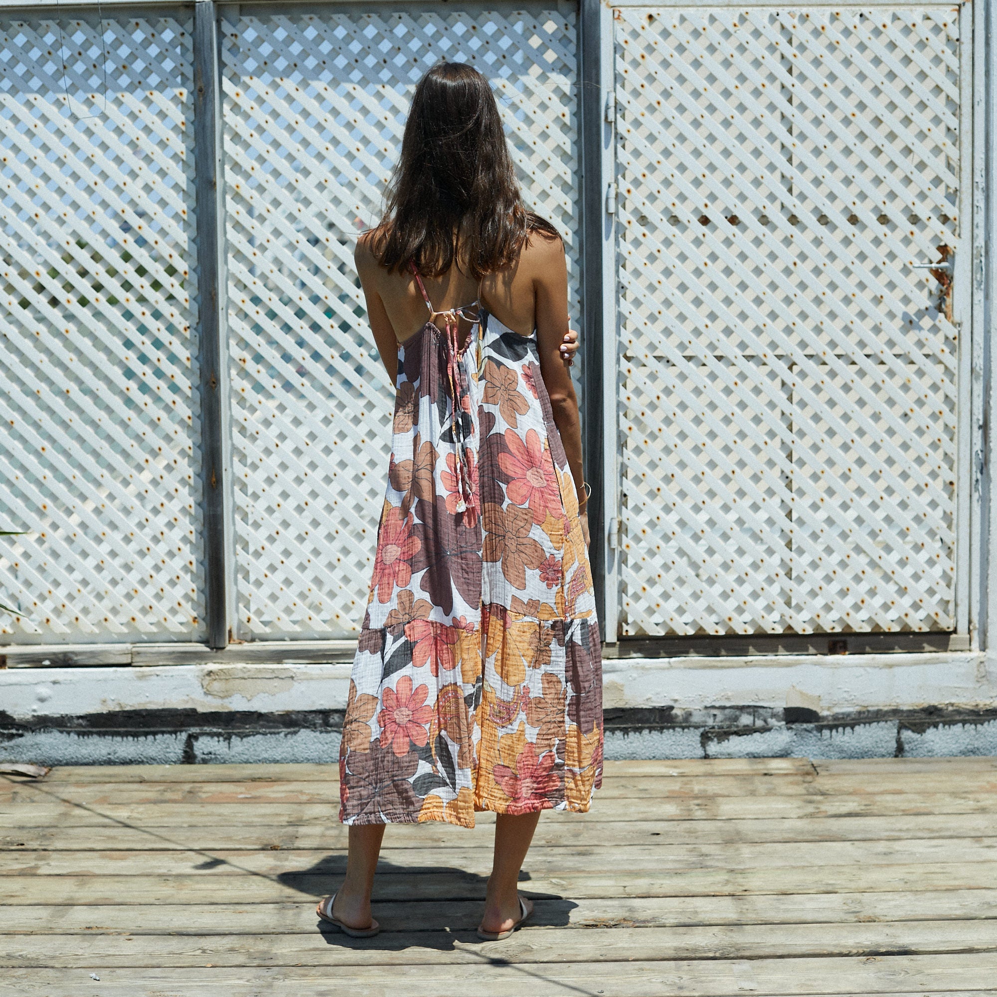 Woman wearing Solara 70s Floral Maxi Dress with orange floral print, standing outdoors against a white lattice screen.