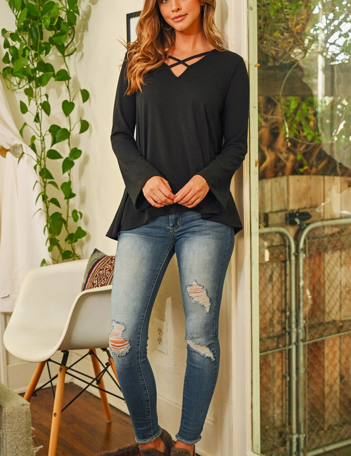 Evanthira Criss-Cross Knit Top with bell sleeves, styled with ripped jeans, showcased in a cozy indoor setting with green plants.