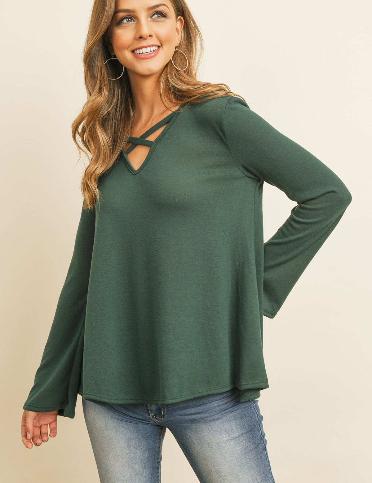 Woman wearing Evanthira Criss-Cross Knit Top in green, featuring a criss-cross neckline and flared bell sleeves, paired with jeans.