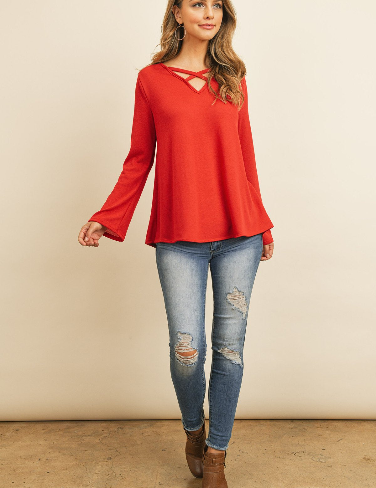 Woman wearing Evanthira Criss-Cross Knit Top in red, paired with ripped jeans and brown boots, featuring bell sleeves and criss-cross neckline.