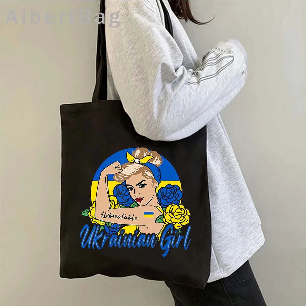 Person holding black tote bag with Ukrainian patriotic design, featuring a strong woman illustration and floral elements.