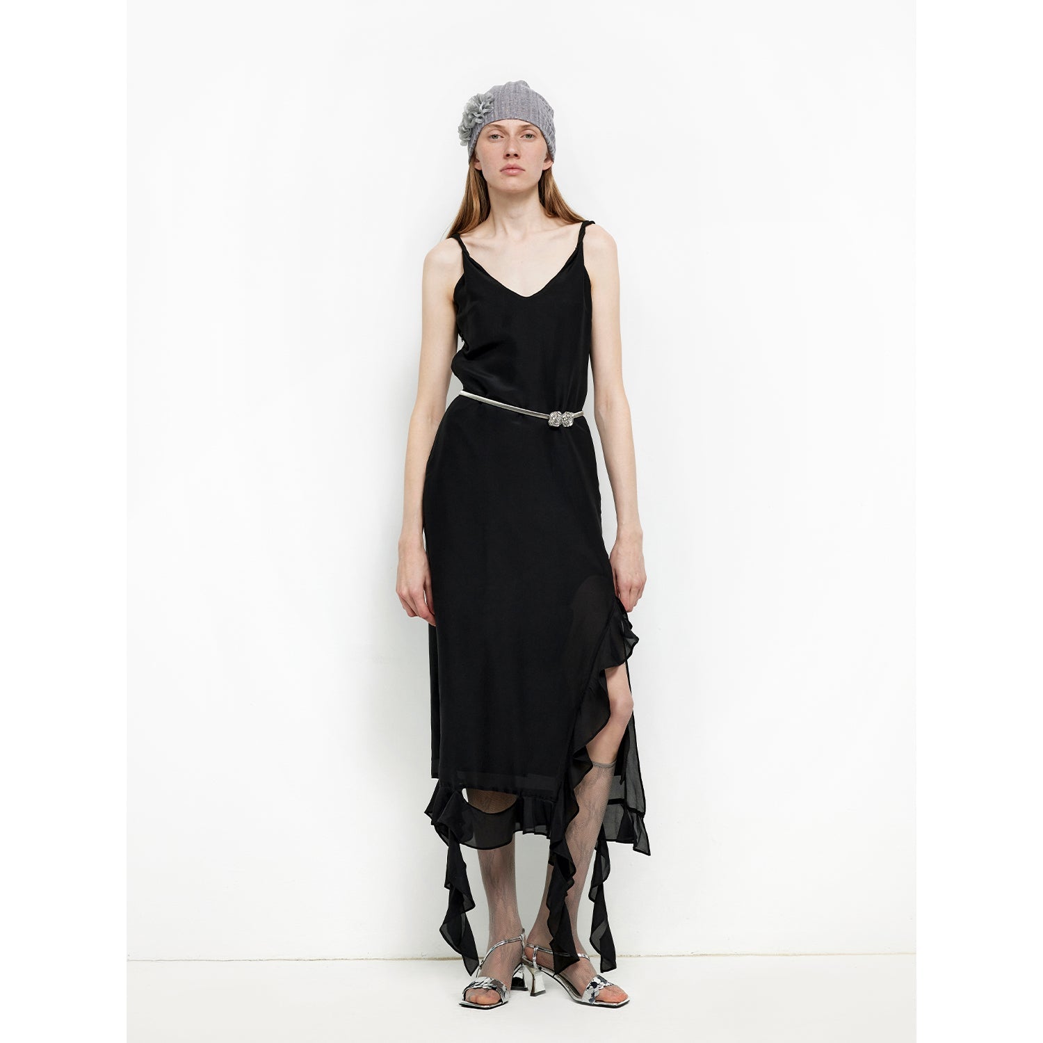Model wearing a black sleeveless dress with asymmetrical hem, gray headband, and metallic sandals, standing against white background.