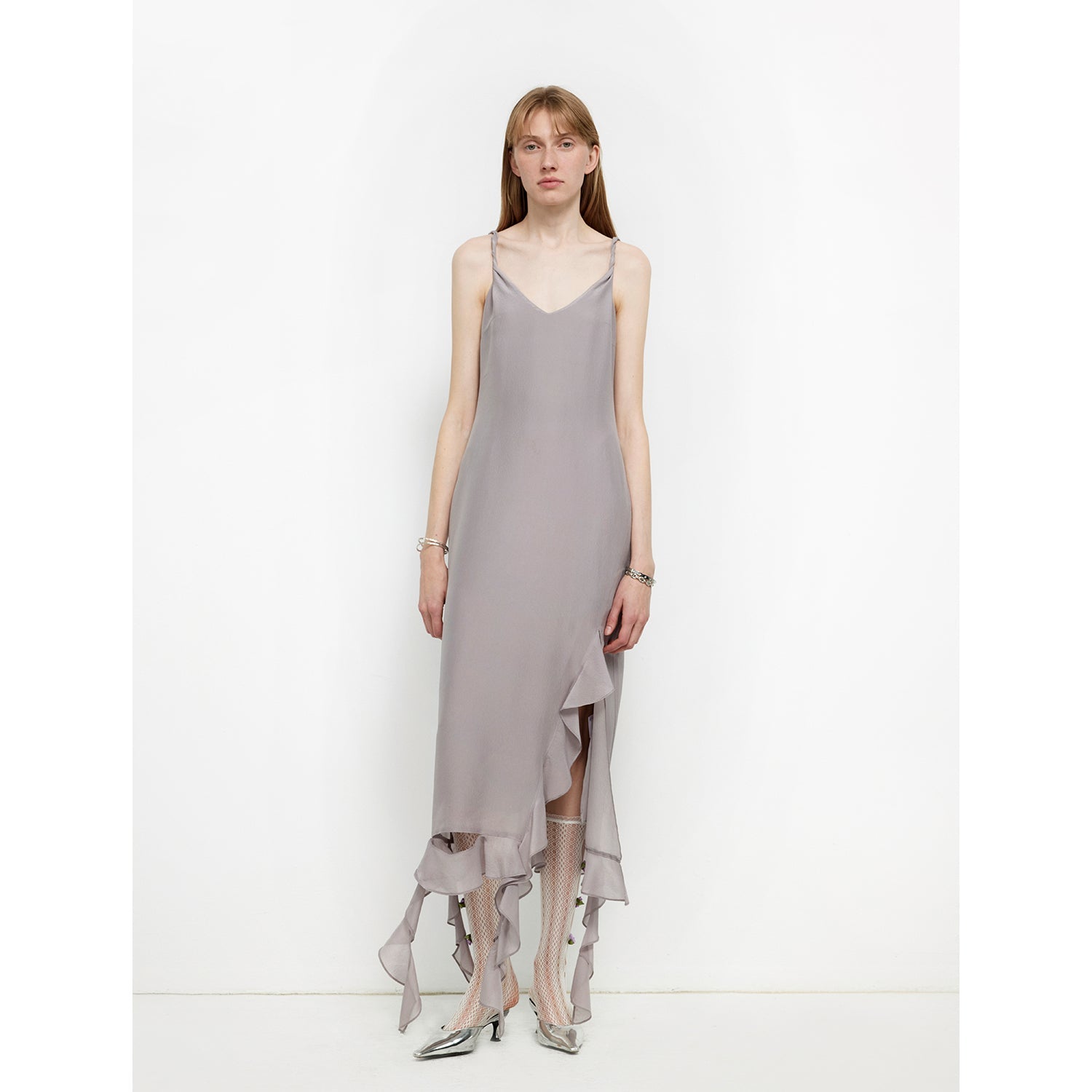 Woman wearing a grey sleeveless dress with a ruffled asymmetrical hem and silver shoes, standing against a white background.