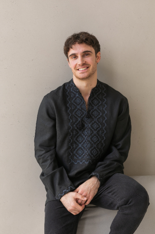 Smiling person wearing a dark embroidered shirt, sitting against a neutral background.