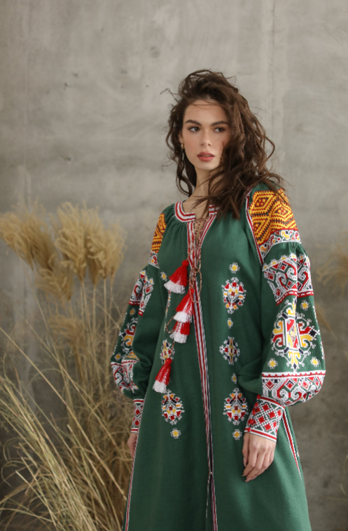 Woman in traditional embroidered dress with colorful patterns, standing by dried plants against a textured wall background.