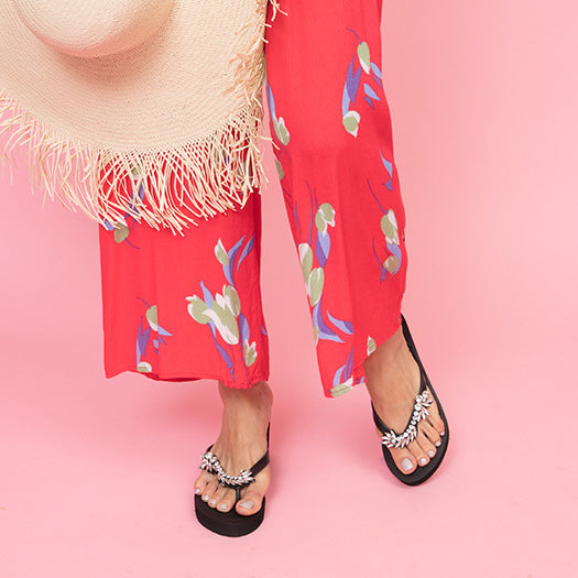 Woman wearing Nomad rhinestone flat flip flop sandals with red floral pants, showcasing crystal vintage motifs on pink background.