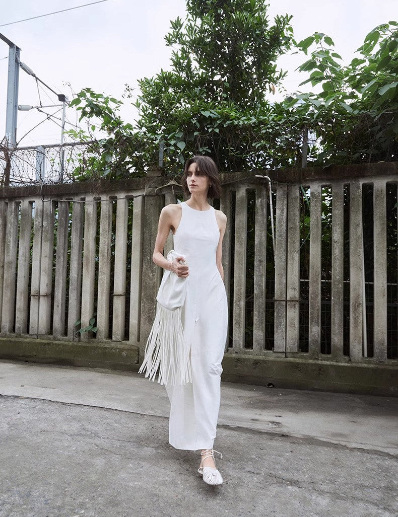 Woman wearing a white sleeveless A-Line dress with a fringed bag, walking beside a wooden fence with lush greenery in the background.
