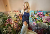 Artist in denim apron holding colorful textured floral paintings