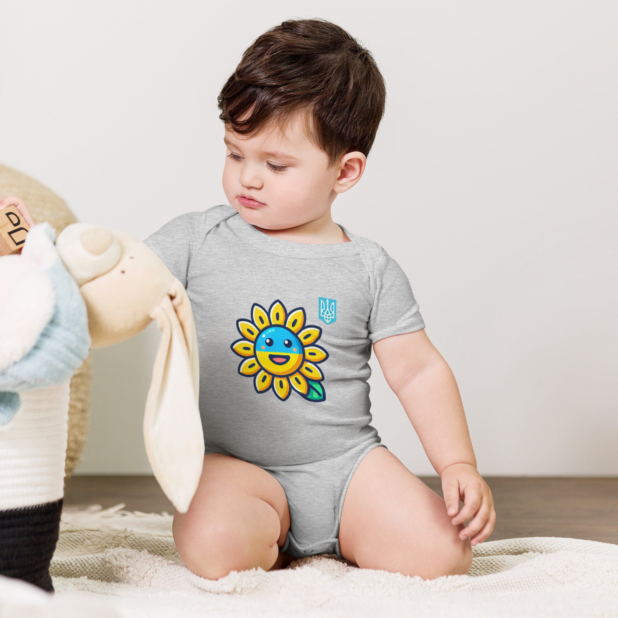 Baby wearing sunflower and Ukrainian flag bodysuit, sitting with stuffed toy.