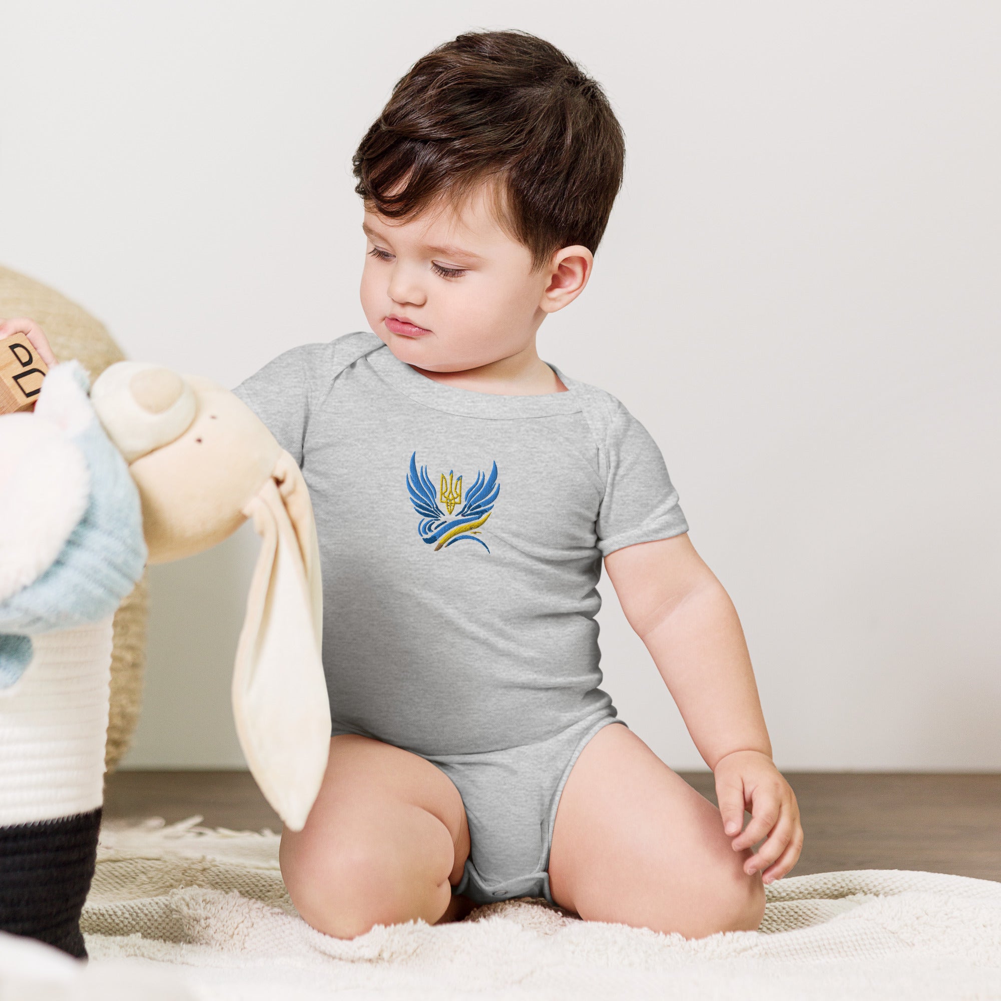 Baby wearing short sleeve one piece with Ukrainian Tryzub logo, sitting on a soft surface.