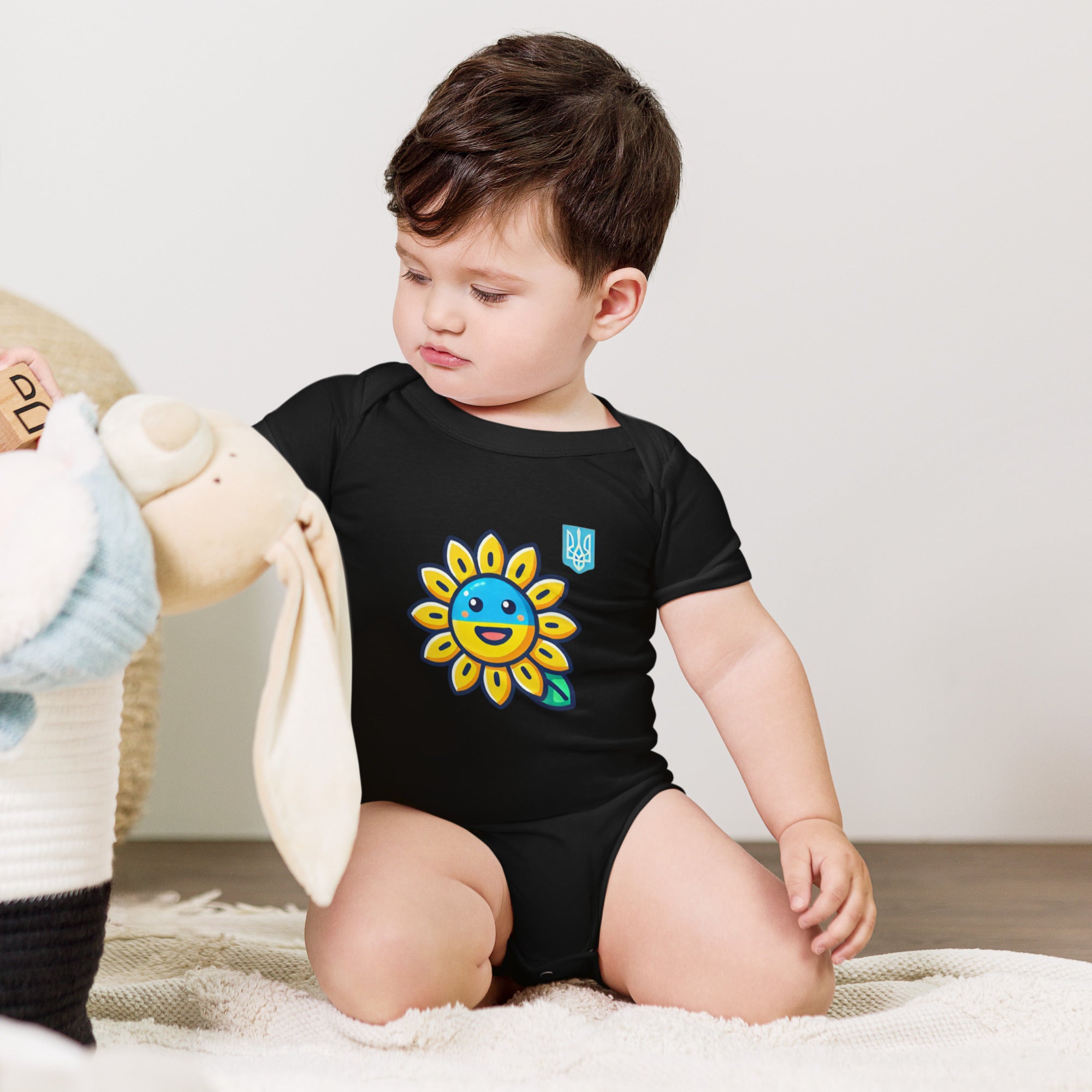 Baby wearing a black bodysuit with sunflower and Ukrainian flag design sitting on a rug with plush toys nearby.