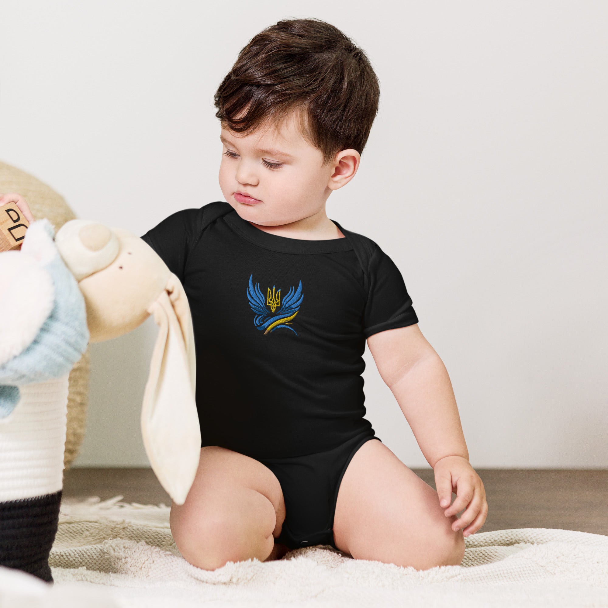 Baby wearing black one-piece romper with Ukrainian Tryzub logo, sitting on a soft rug and playing with a plush toy.