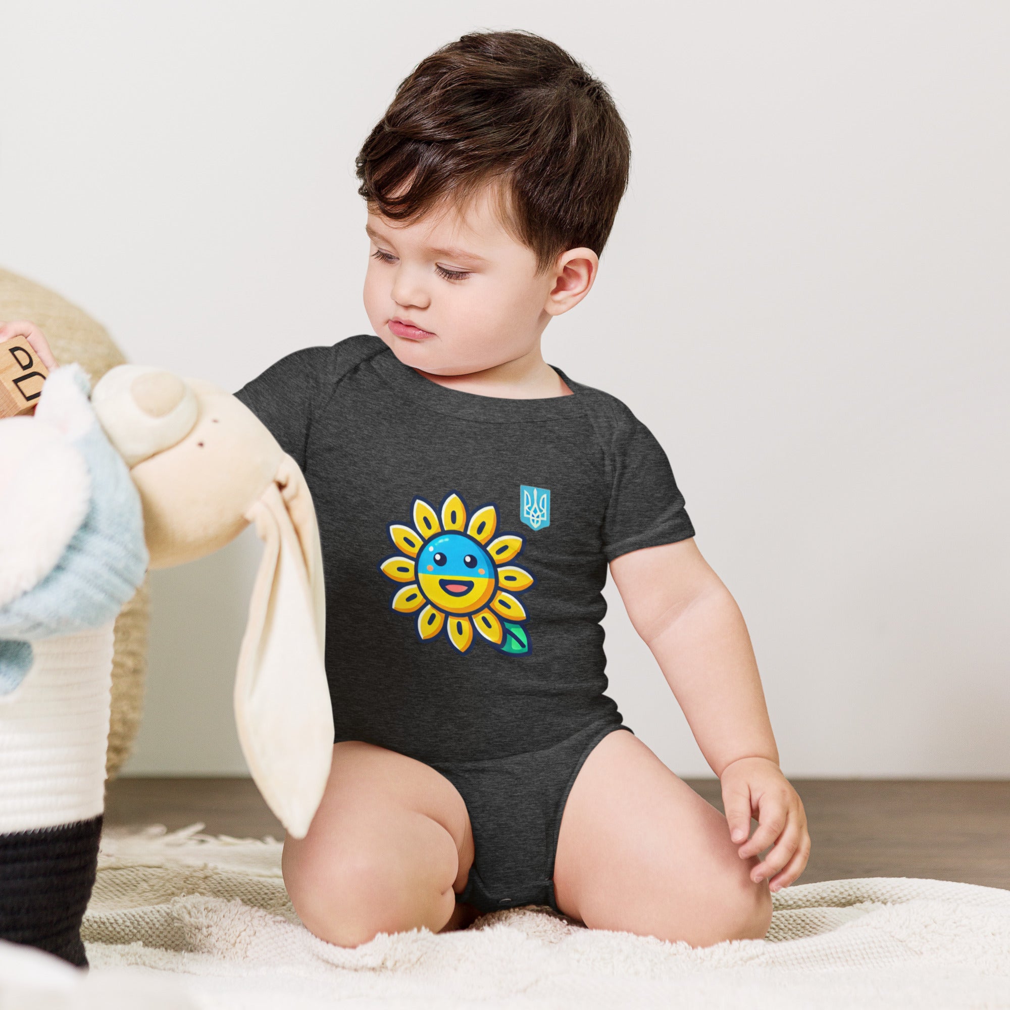 Baby wearing sunflower and Ukrainian flag bodysuit, sitting with stuffed toy.