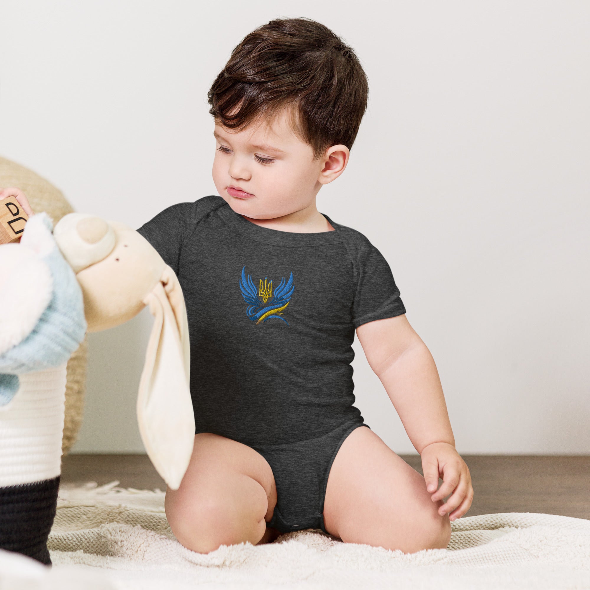 Baby wearing short sleeve one piece with Ukrainian Tryzub logo, sitting on a soft rug next to a plush toy.