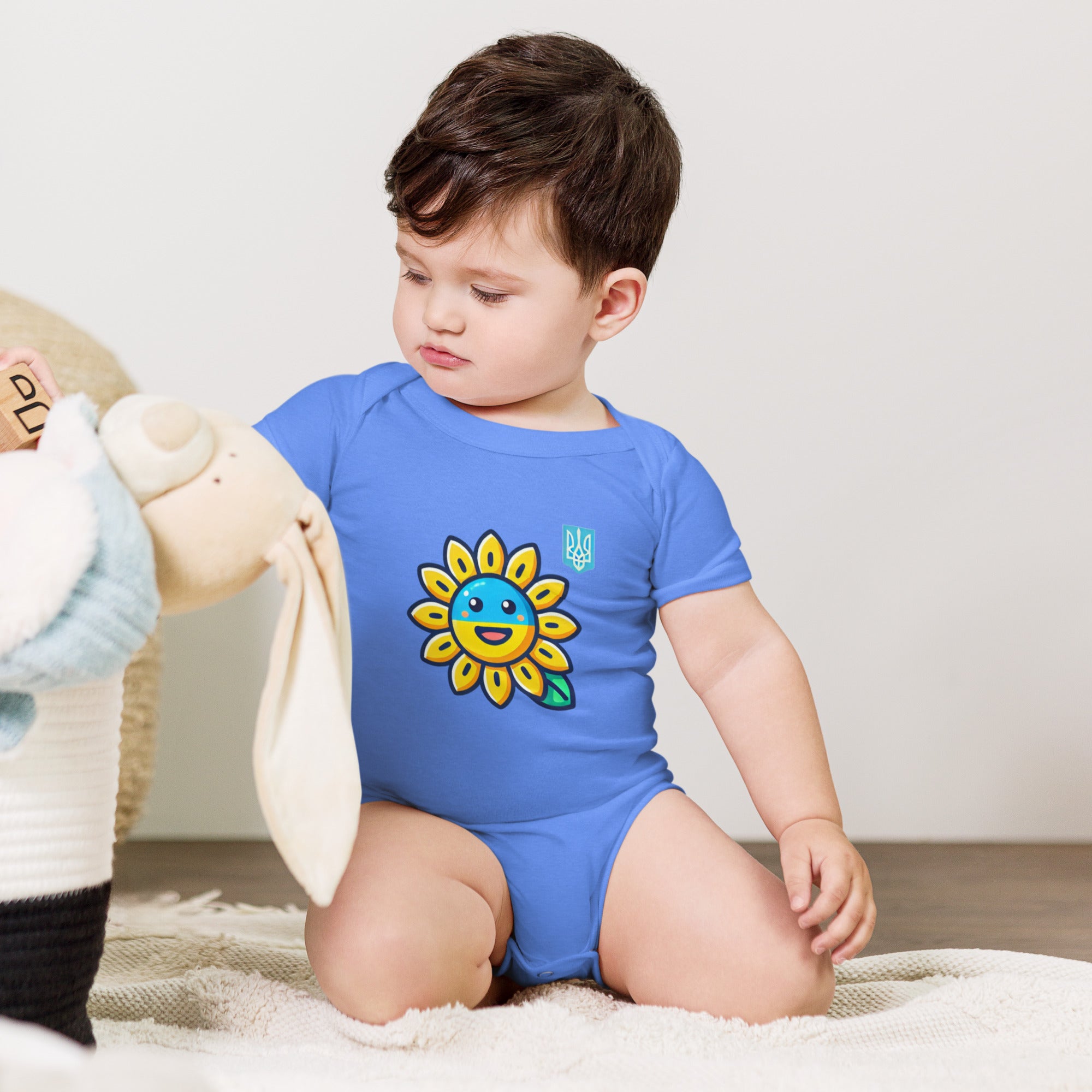 Baby wearing a blue bodysuit with sunflower and Ukrainian flag design, sitting on a soft surface with a plush toy nearby.